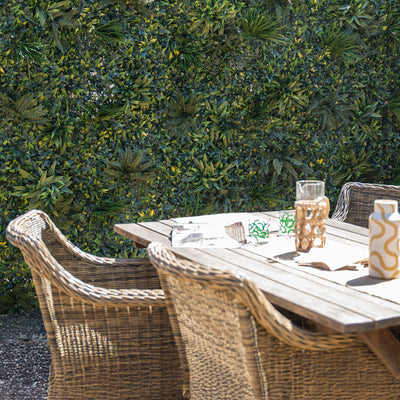 Outdoor setting with a wooden table and wicker chairs against a green foliage wall.