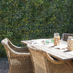 Outdoor setting with a wooden table and wicker chairs against a green foliage wall.