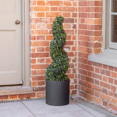 An artificial tall, spiral-shaped green topiary tree in a matte black cylindrical planter, placed on a stone patio corner against a red brick wall and a beige door.