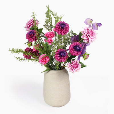 A faux floral arrangement featuring pink, purple, and magenta flowers with green foliage, presented in a textured light beige ceramic vase against a clean white background.