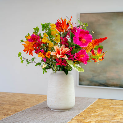 An artificial flower bouquet full of vibrant and exotic flowers in a textured off-white ceramic vase, displayed on a wooden table runner in front of a neutral wall and framed artwork.