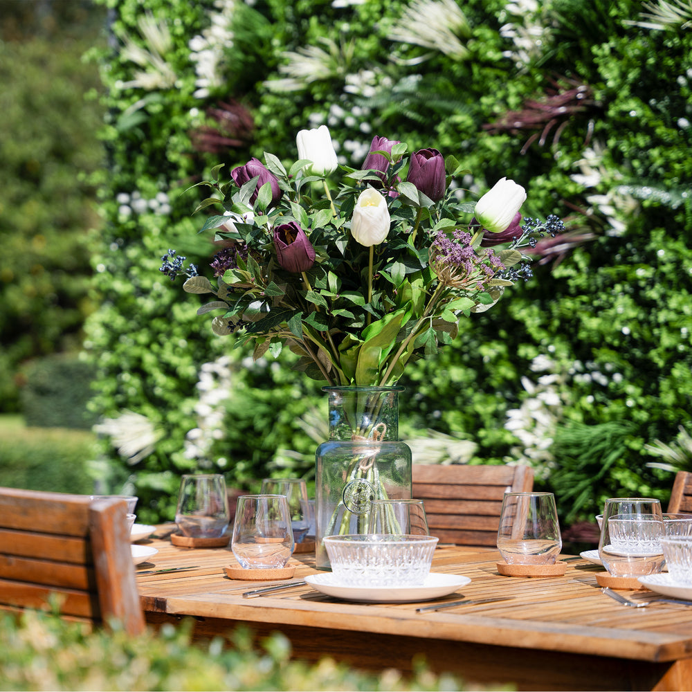 Bouquet of purple and white flowers in a clear vase on a wooden table outdoors.