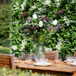 Bouquet of purple and white flowers in a clear vase on a wooden table outdoors.