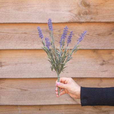 A hand holding a small bouquet of artificial lavender stems against a rustic wooden plank wall.