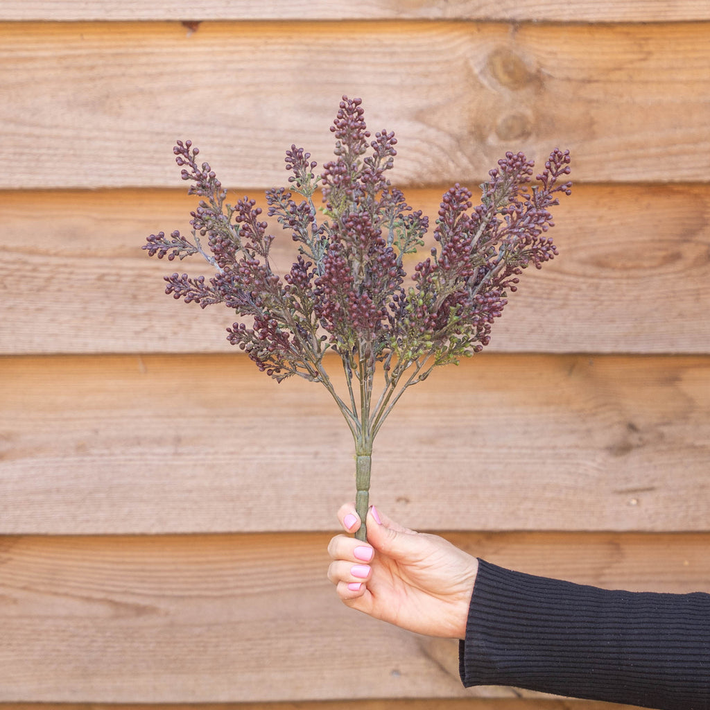 A hand holding a small bouquet of artificial burgundy-purple berries on a spike against a background of wooden panelling.