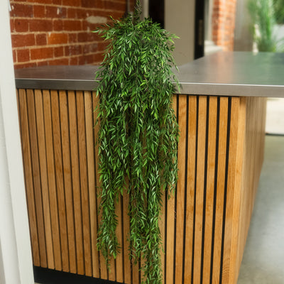 A green trailing houseplant with dense leafy foliage draped over a modern wooden slat counter in an indoor setting with a brick wall and shelving in the background.