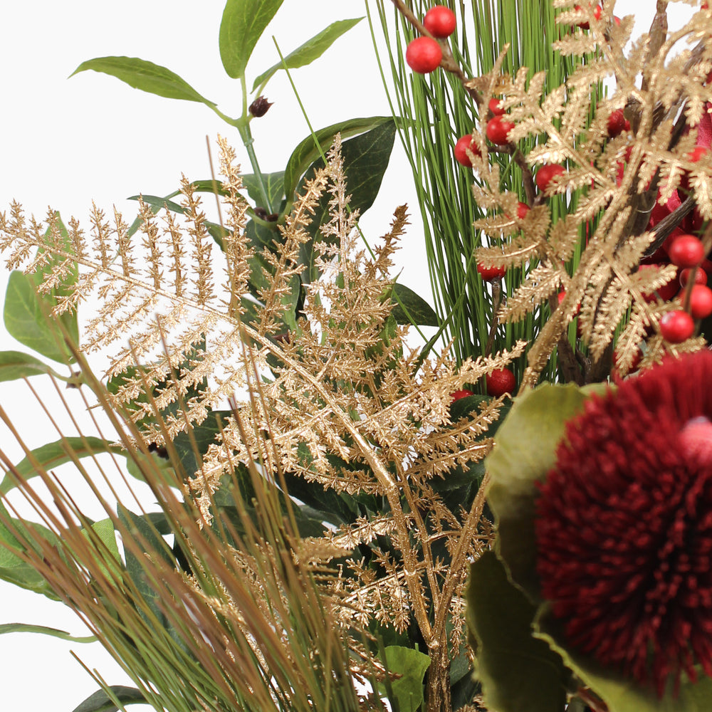 A close-up of an artificial floral arrangement featuring gold ferns, bay leaves, small red berries, and a prominent deep red thistle head on a white background.