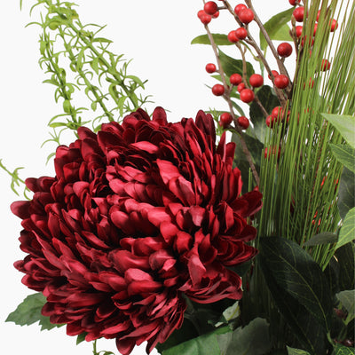 A close-up of an artificial floral arrangement featuring a large deep red chrysanthemum bloom, pine and bay foliage, and clusters of small red berries on a white background.