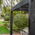 Two spherical green artificial topiary bushes hanging from a black metal frame on a pergola in a backyard garden setting.