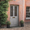 A gray front door with decorative stained-glass panels, set in a red brick entryway with ivy on the left, a window on the right, and two round green topiary bushes in black planters on the gravel ground.