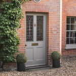 A gray front door with decorative stained-glass panels, set in a red brick entryway with ivy on the left, a window on the right, and two round green topiary bushes in black planters on the gravel ground.