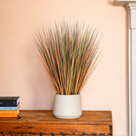 A decorative artificial grass arrangement in a textured off-white ceramic pot, displayed on a rustic wooden chest against a soft peach-pink wall.