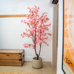 Pink artificial cherry blossom tree in a pot next to a wooden chest against a white wall.