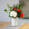 An artificial floral arrangement in a textured off-white ceramic vase, featuring white hydrangeas, red peonies, and holly and berries on a table runner.