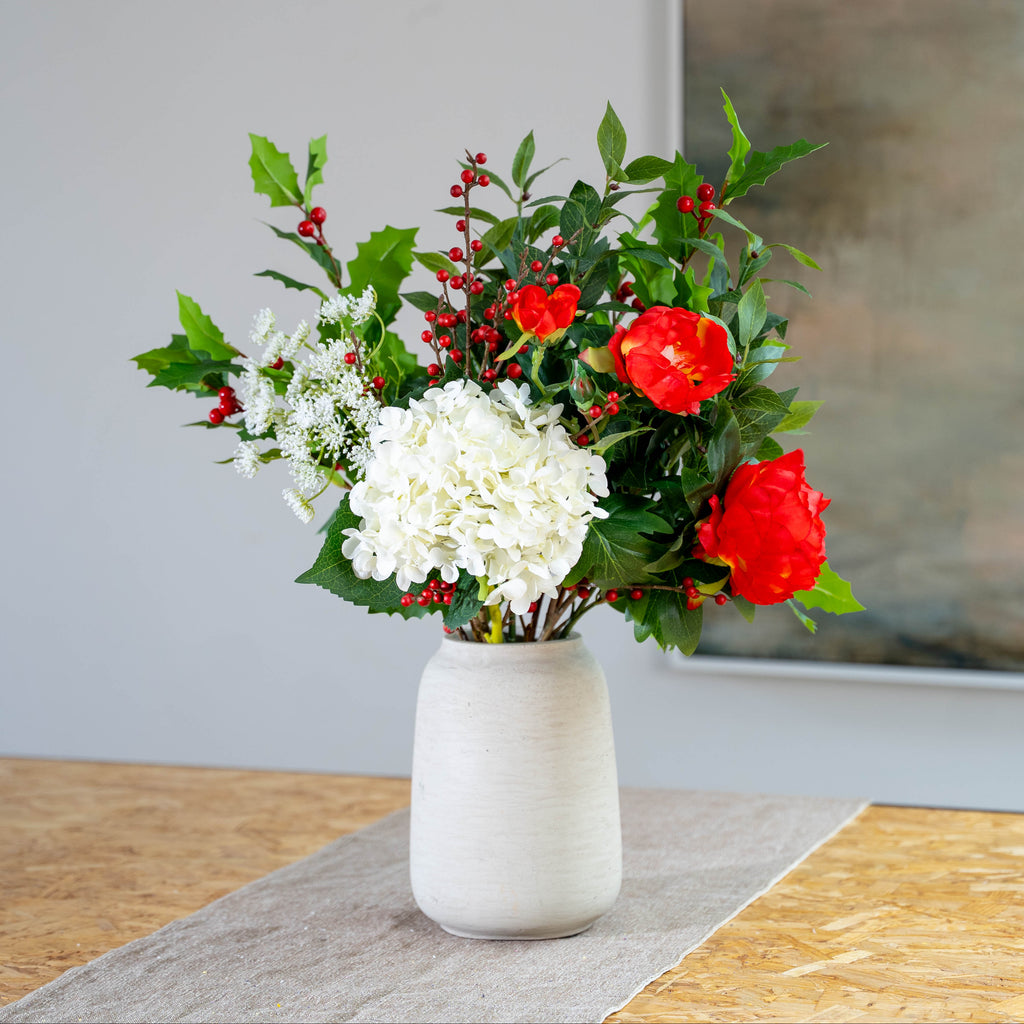 An artificial floral arrangement in a textured off-white ceramic vase, featuring white hydrangeas, red peonies, and holly and berries on a table runner.