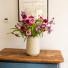 A vibrant floral arrangement featuring pink, purple, and red flowers with green foliage, presented in a textured cream-colored ceramic vase on a rustic wooden table.
