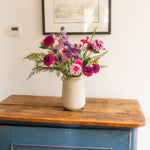 A vase of mixed artificial flowers in pink, purple, and magenta tones with green foliage, displayed on a rustic wooden cabinet against a white wall.