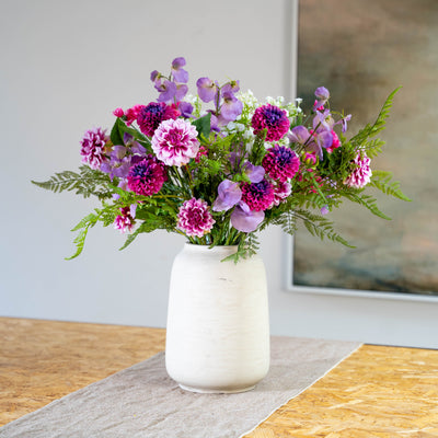 A vibrant, purple, pink and white artificial flower bouquet with woodland foliage in a textured off-white ceramic vase, placed on a neutral runner on a wooden table with a blurred interior background.