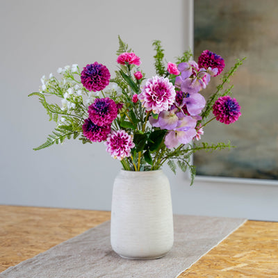 A vibrant, purple, pink and white, artificial flower arrangement with woodland foliage in a textured off-white ceramic vase, set on a wooden table with a neutral background.