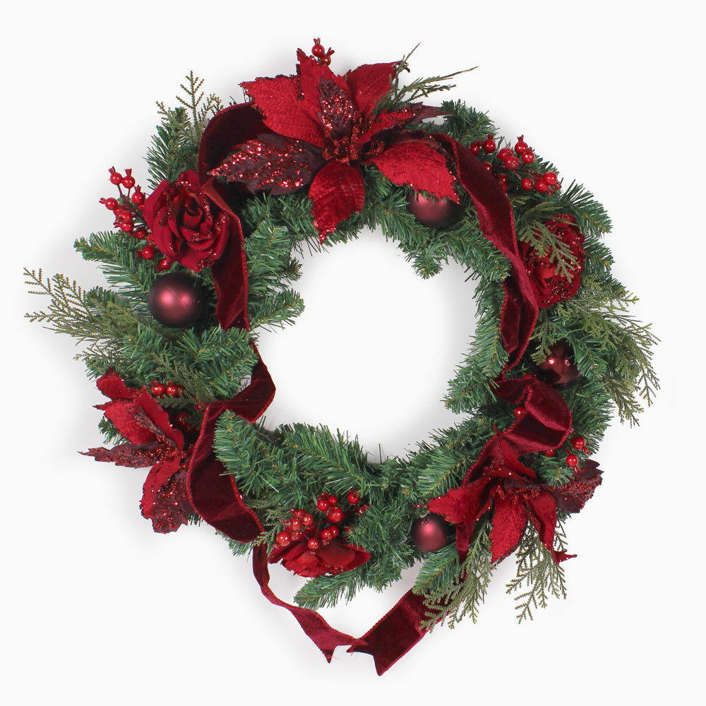 A festive Christmas wreath decorated with green pine branches, red glitter poinsettias, red velvet ribbons, red berries, and dark red ornaments, shown against a plain white background.