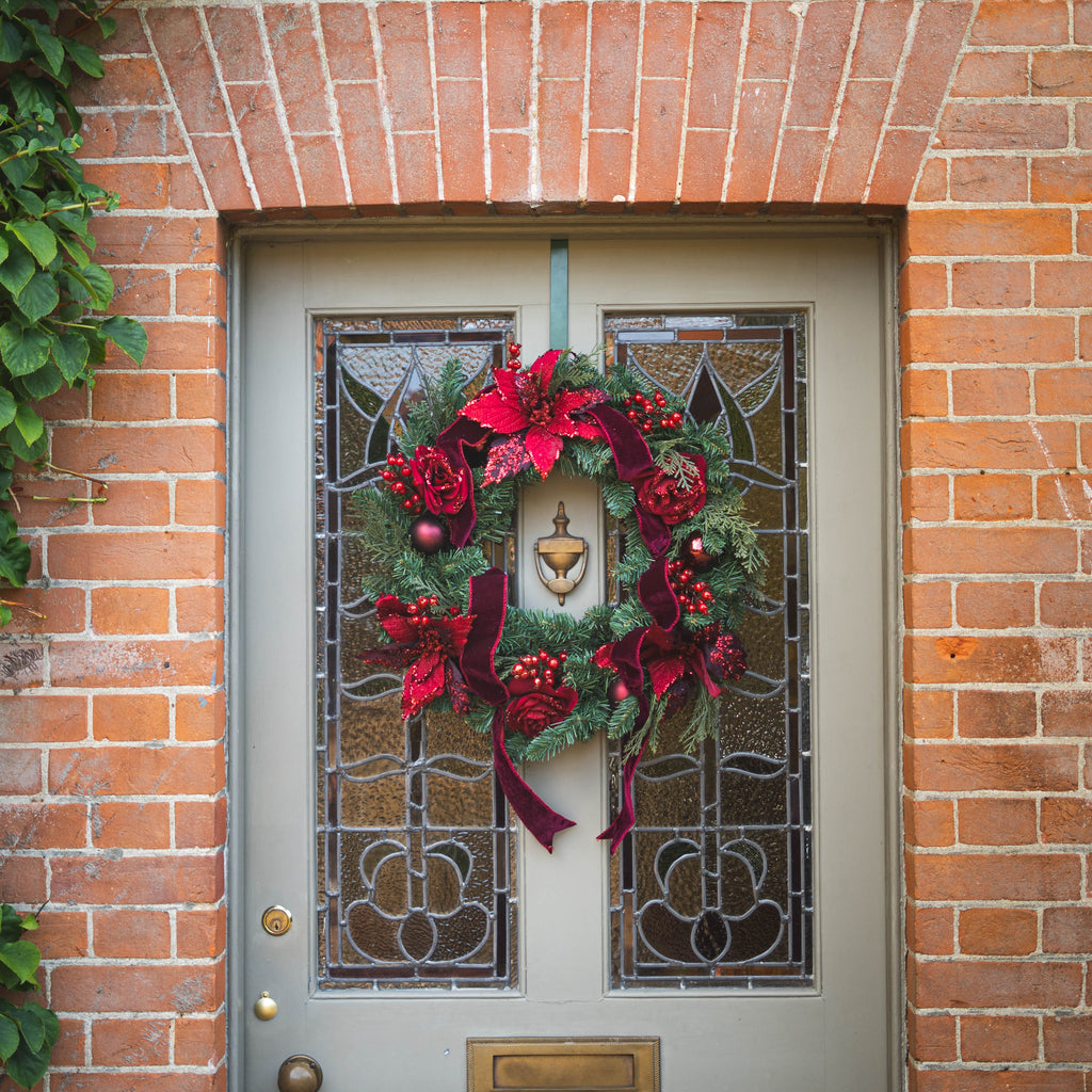 A festive Christmas wreath with red poinsettias, burgundy ribbons, green pine branches, and red berries hangs on a gray front door with decorative stained glass panels, set within a brick wall.