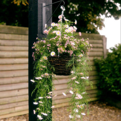 A hanging basket filled with trailing pink and white flowers, suspended from a black metal bracket on a dark post in a garden setting with a wooden fence in the background.
