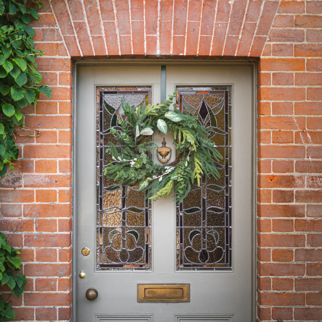 A green holiday wreath with pine branches displayed on a gray front door with decorative stained glass panels, set within a red brick wall.
