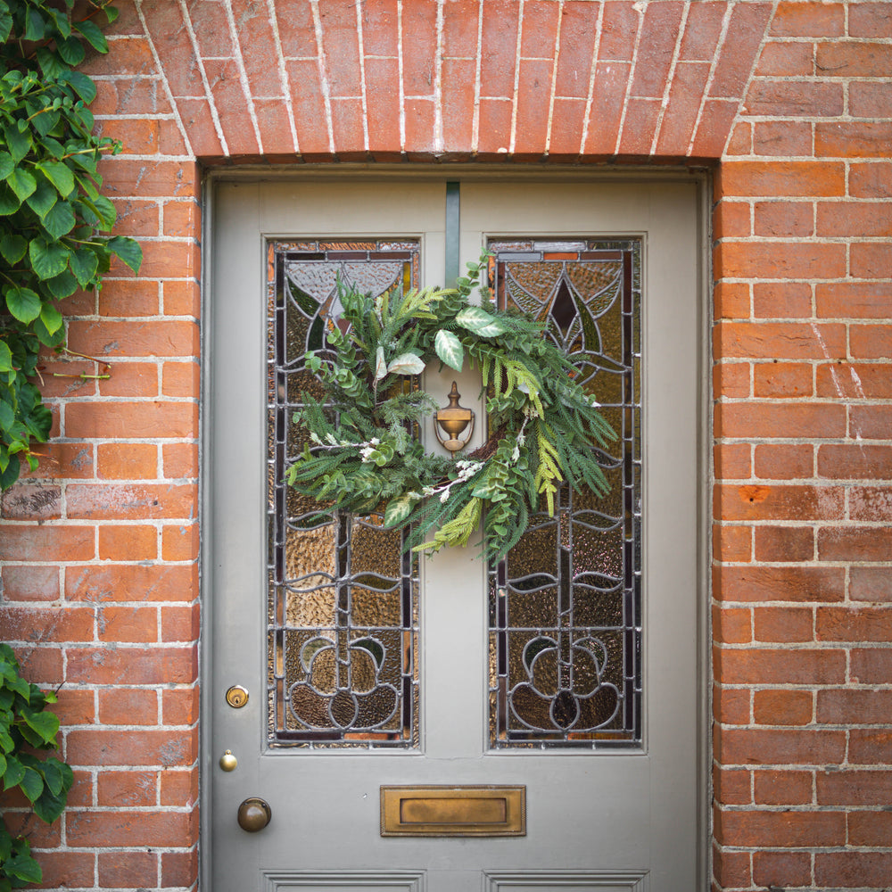 A green holiday wreath with pine branches displayed on a gray front door with decorative stained glass panels, set within a red brick wall.