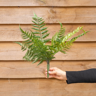 A green artificial fern branch on a planting spike held by a hand against a brown wooden plank wall background.