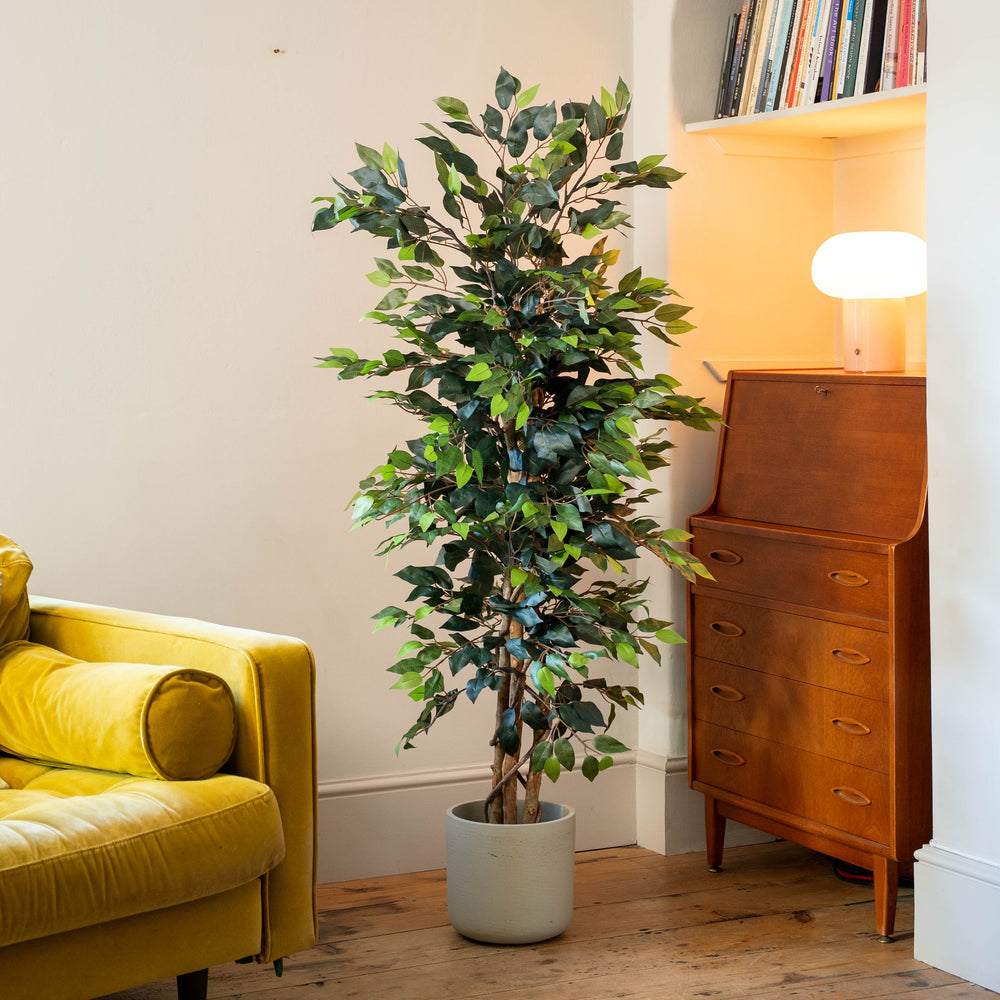 A tall artificial potted tree with green leaves stands in a corner of a room next to a yellow armchair and a mid-century modern wooden cabinet.