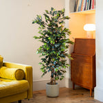 A tall artificial potted tree with green leaves stands in a corner of a room next to a yellow armchair and a mid-century modern wooden cabinet.