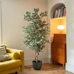 A tall, leafy potted artificial tree with variegated green and cream leaves stands in a dark gray pot in a cozy living room corner next to a yellow sofa and a mid-century modern wooden cabinet.
