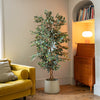 A tall artificial variegated ficus tree in a white concrete pot, styled in a cozy living room corner next to a yellow sofa and a wooden cabinet.