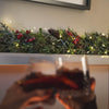 A festive holiday scene with a green garland decorated with pinecones, red berries, and warm string lights across a shelf, while two blurred hands in the foreground hold up glasses of red wine.