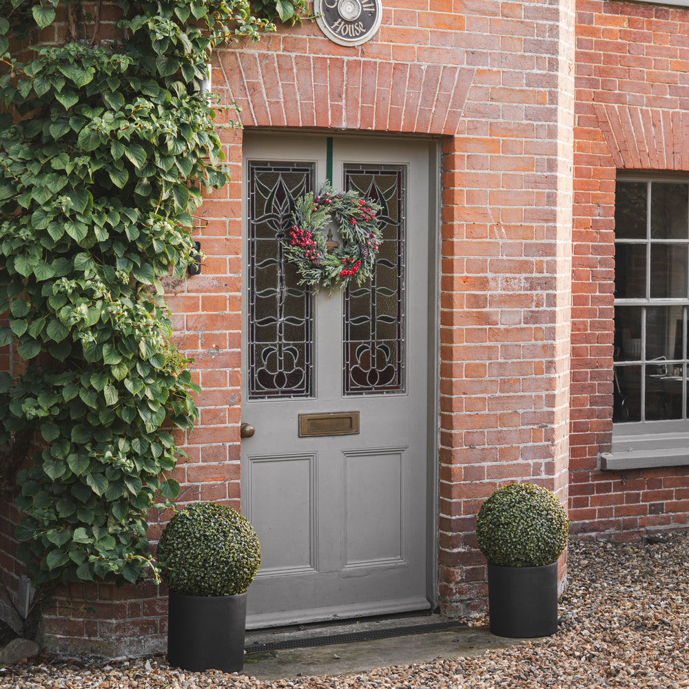 A gray front door with decorative glass panels, adorned with a red berry wreath, set in a brick entryway with ivy, potted topiary trees, and a house number plaque.