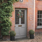 A gray front door with decorative glass panels, adorned with a red berry wreath, set in a brick entryway with ivy, potted topiary trees, and a house number plaque.