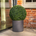 A spherical green topiary bush in a matte dark gray cylindrical planter, set against a brick wall and glass door.