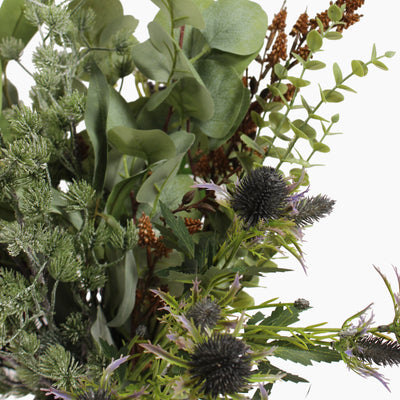 An artificial floral arrangement featuring soft green eucalyptus leaves, thistle stems with dark spiky blooms, and frosted green pine foliage, photographed against a plain white background.