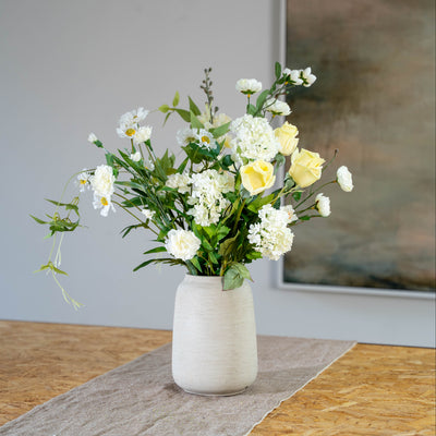 A faux flower arrangement featuring white, cream, and pale yellow flowers with green trailing foliage, displayed in a textured off-white ceramic vase on a grey table runner.