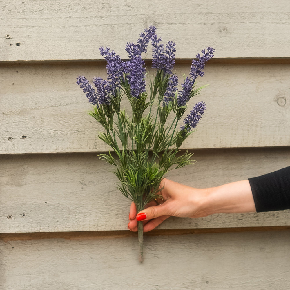A hand with red nail polish holds a small bunch of artificial lavender stems with purple flowers and green leaves against a light gray wooden plank background.