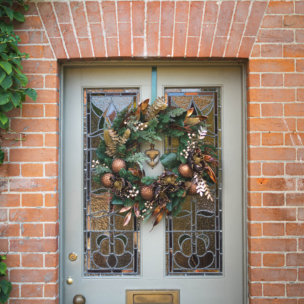 An autumn-themed Christmas wreath with copper ornaments, pinecones, and evergreen branches hanging on a gray front door with stained glass panels, set in a brick doorway.