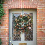 An autumn-themed Christmas wreath with copper ornaments, pinecones, and evergreen branches hanging on a gray front door with stained glass panels, set in a brick doorway.