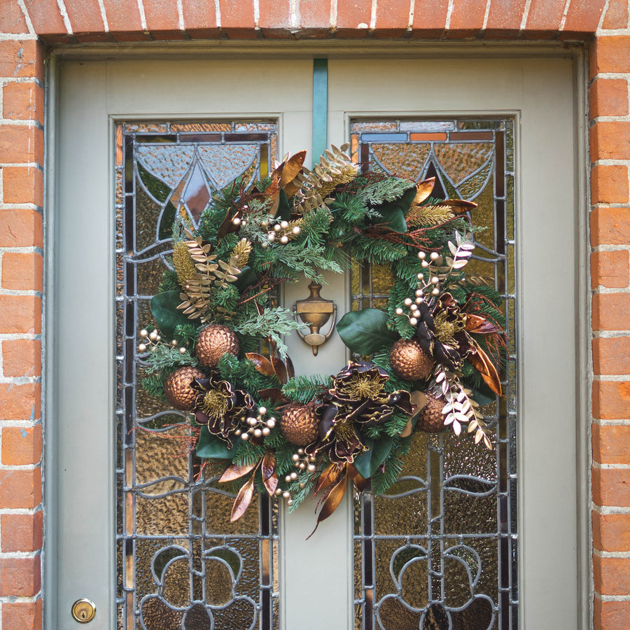 An autumn-themed Christmas wreath with copper ornaments, pinecones, and evergreen branches hanging on a gray front door with stained glass panels, set in a brick doorway.