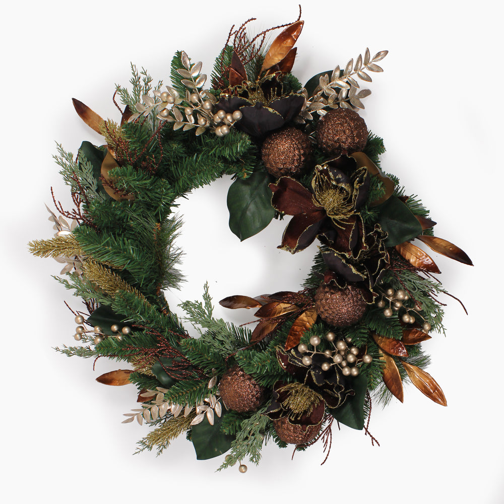 An artificial holiday wreath featuring green pine branches, dark green magnolia leaves, bronze-brown pinecones, gold-toned accents, and small gold berry clusters, photographed against a plain white background.