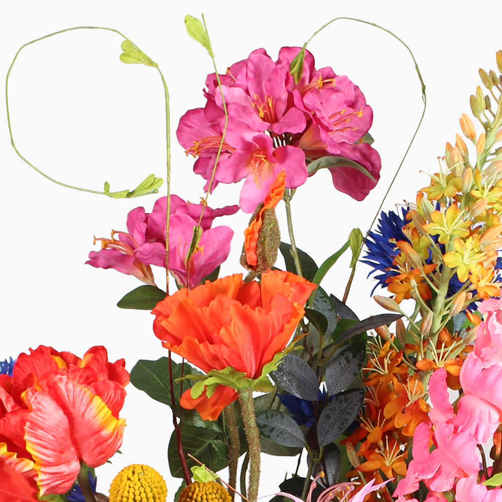 A close-up of a mixed floral arrangement featuring bright pink, orange, red, yellow, and blue flowers with tall spiralling tendrills on a plain white background.