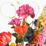 A close-up of a mixed floral arrangement featuring bright pink, orange, red, yellow, and blue flowers with tall spiralling tendrills on a plain white background.