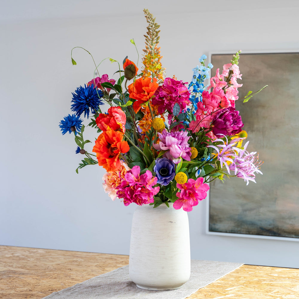 A vibrant, multicolored artificial flower bouquet arranged in a textured white ceramic vase, set on a wooden table against a neutral wall with a framed painting.