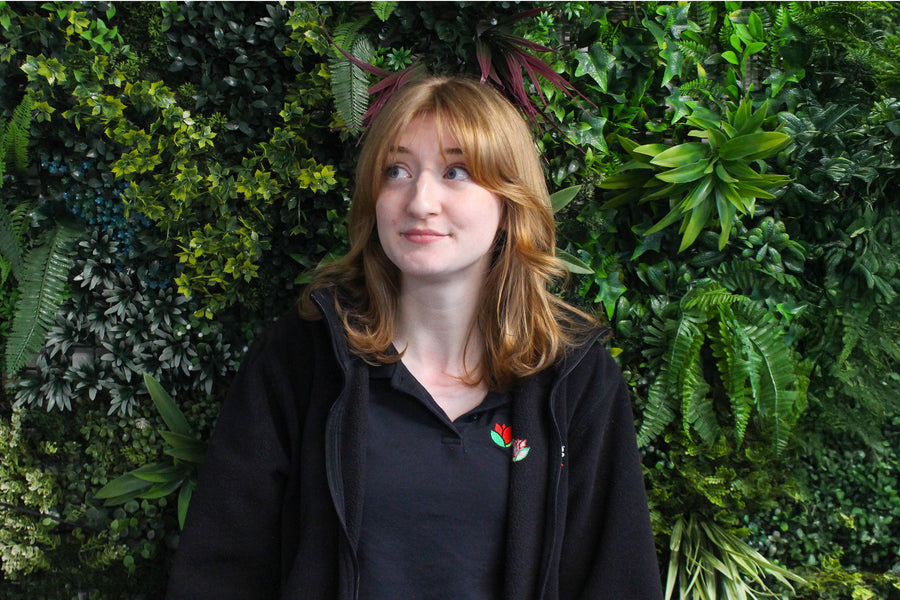 Melissa, a member of staff, wearing a black uniform in front of a green leafy wall.