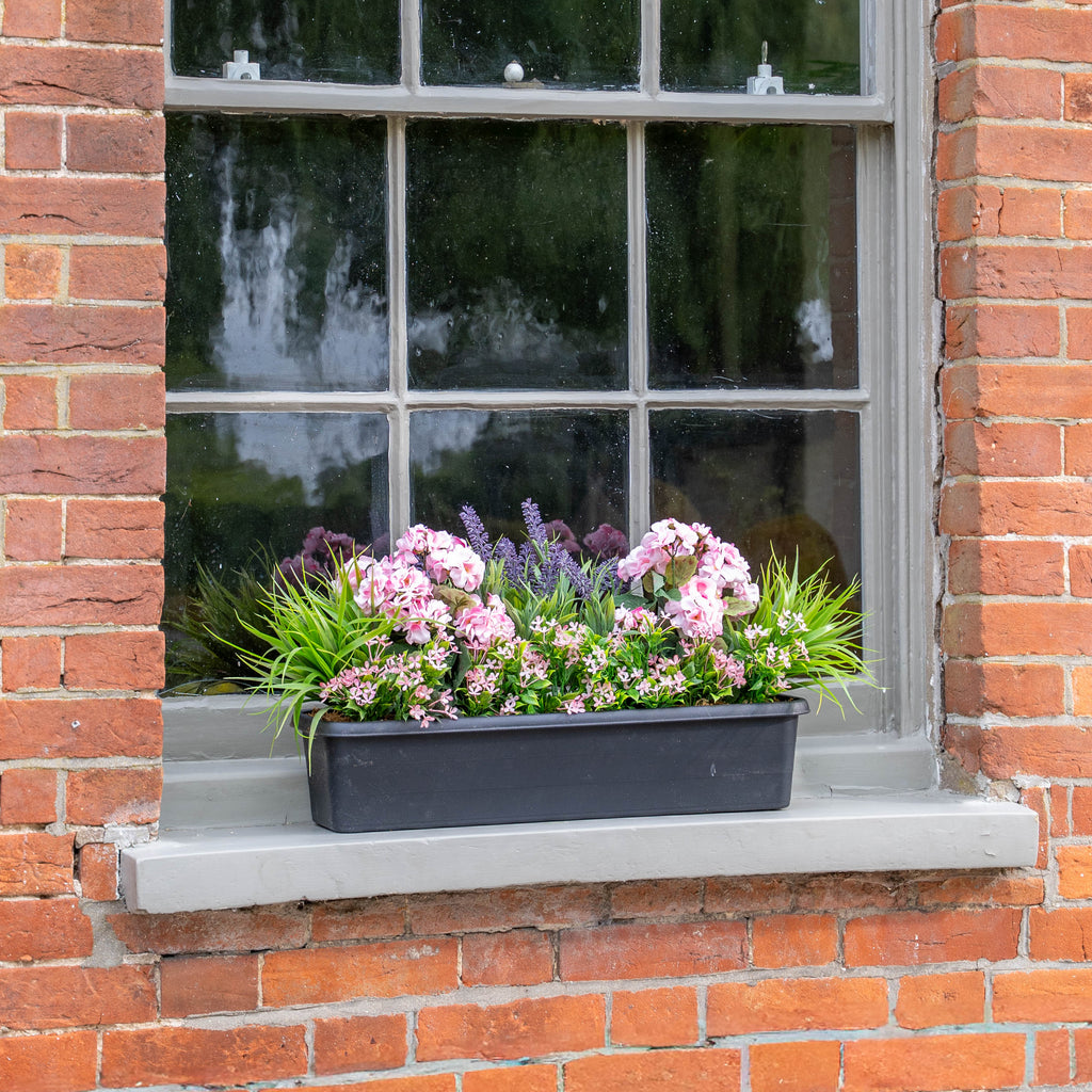 A rectangular black window box planter filled with pink and purple flowers and green foliage, displayed on a gray stone window sill of a red brick building.