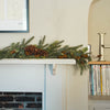 A green pine garland with brown pinecones and warm string lights draped across a white fireplace mantel in a home interior setting.